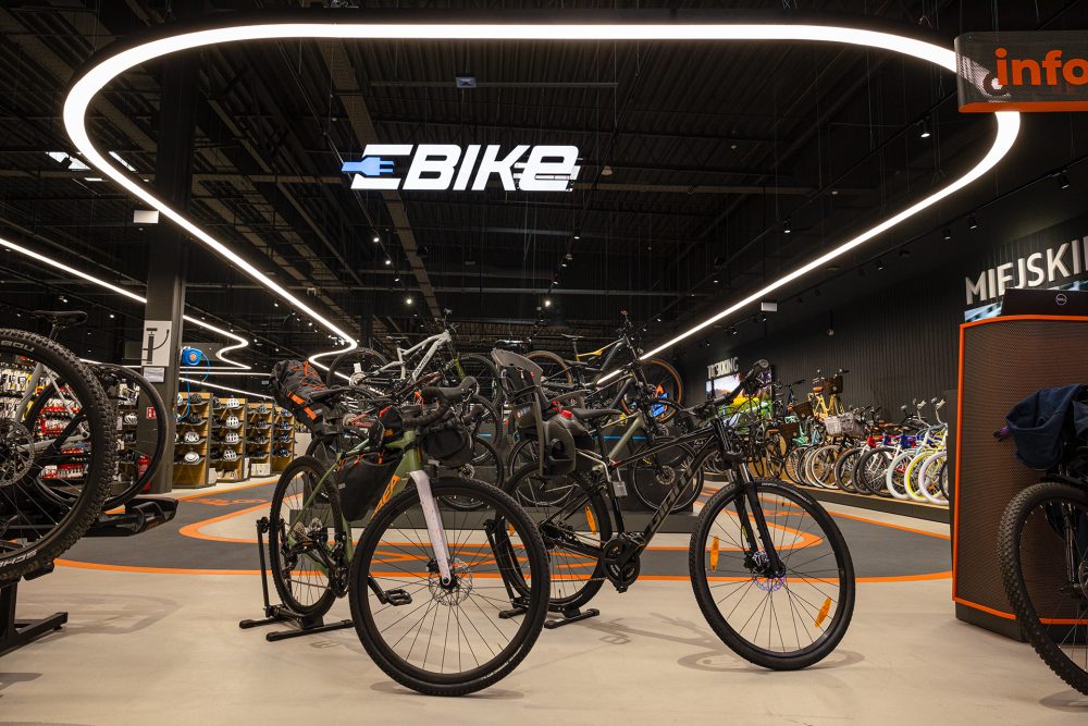 Wnętrze nowoczesnego salonu rowerowego z ekspozycją rowerów miejskich, trekkingowych i elektrycznych ustawionych na stojakach pod charakterystycznym oświetleniem sufitowym.